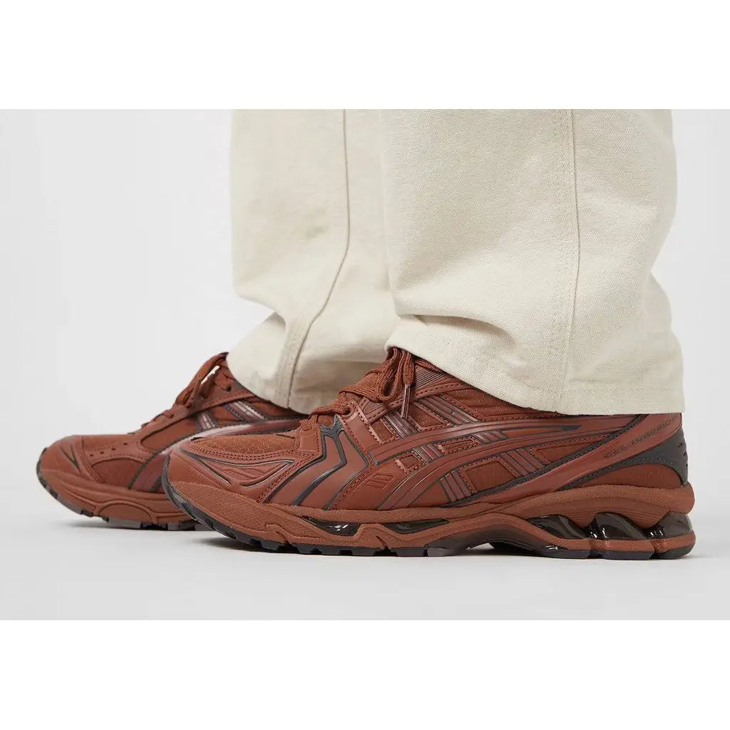 Brown athletic shoes with black accents worn by a person in beige pants on a white background